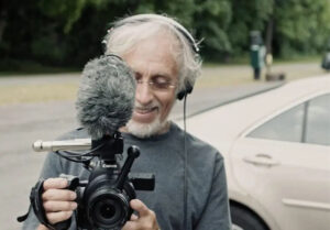 Older man stands in front of a car with camera equipment